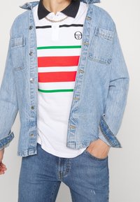 Světlá modrá džínová bunda přes bílou polokošili s červenými, zelenými a černými pruhy. Má malé logo a klasický límec. Celkový vzhled doplňují přiléhavé džíny.