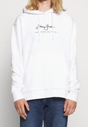 Sweatshirt blanc avec capuche à cordon de serrage et grande poche avant. Présente un logo de marque en cursif noir et des noms de lieux imprimés au centre.