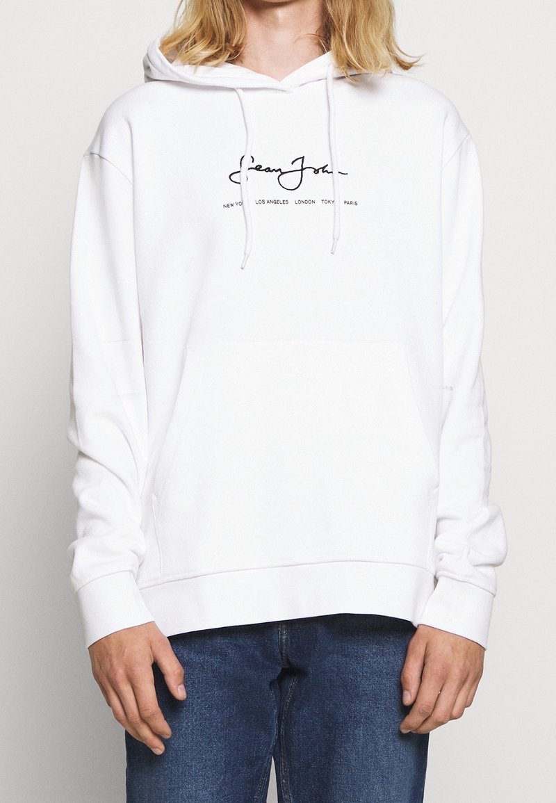 Sweatshirt blanc avec capuche à cordon de serrage et grande poche avant. Présente un logo de marque en cursif noir et des noms de lieux imprimés au centre.