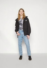Svart vanntett jakke med hette, brukt over en grafisk t-skjorte, kombinert med lyseblå denimjeans og svart fottøy. Minimalistisk design.