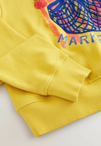 Gele sweatshirt met geribbelde manchetten, voorzien van een blauw grafisch ontwerp en rode accenten. Gemaakt van zachte stof met een gestructureerd patroon.