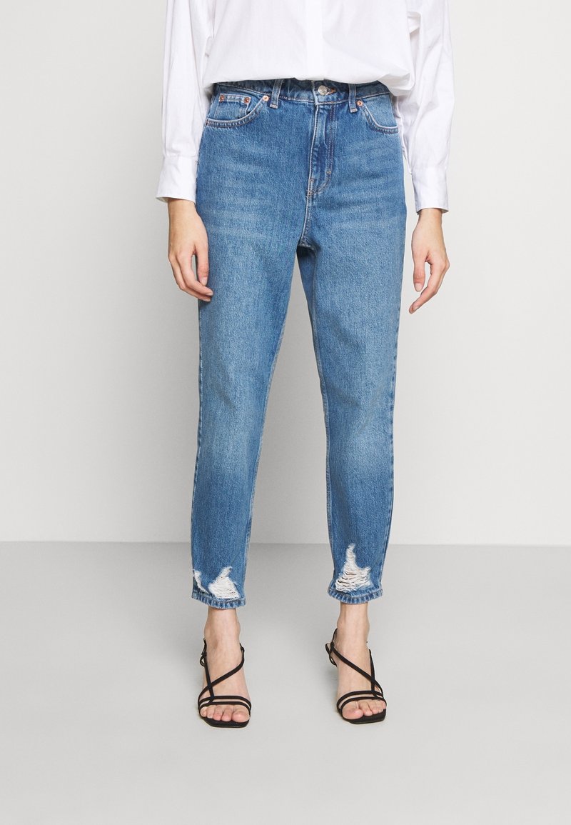 Žena v modrých roztrhaných džínách s kotníkovými manžetami, bílé košili s dlouhým rukávem a černých sandálech na podpatku s otevřenou špičkou stojí na šedé podlaze.