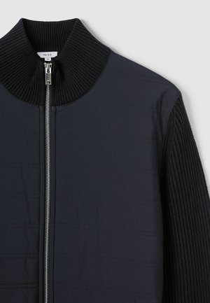 Veste bleu marine avec un col montant à côtes, dotée d'une fermeture éclair sur le devant et d'un tissu texturé avec des lignes horizontales, mettant en valeur différents matériaux.
