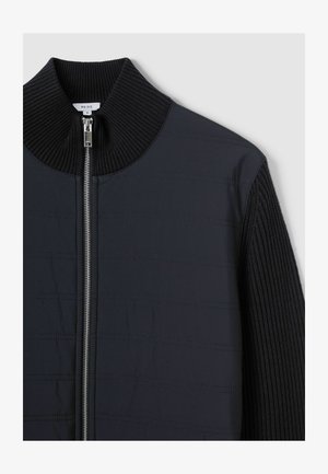Marinejacke mit hohem Rippkragen, verfügt über einen Frontreißverschluss und ein strukturiertes Gewebe mit horizontalen Linien, das verschiedene Materialien hervorhebt.