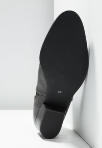 Semelle d'une botte en cuir noir avec un talon bloc, taille 37 imprimée sur la semelle, positionnée contre un mur d'angle blanc.