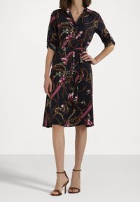 Svart klänning med blom- och kedjemönster, skjortkrage, korta uppvikta ärmar, midjeband, flytande kjol, kombinerad med bruna sandaler med remmar.