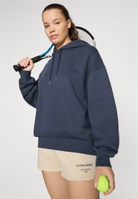 Felpa con cappuccio blu navy dalla vestibilità rilassata, abbinata a pantaloni corti beige con testo nero. Tiene in mano una pallina da tennis verde e una racchetta.