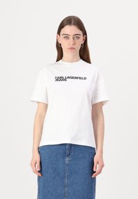 Biele tričko z bavlny s krátkymi rukávmi, s čiernym nápisom "KARL LAGERFELD JEANS" na prednej strane. V kombinácii s džínsovou sukňou.