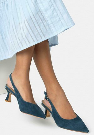 Picioare purtând pantofi cu toc bloc de înălțime medie, culoare teal, din piele întoarsă, cu barete pe călcâi și vârf ascuțit, asortați cu o fustă texturată albastru deschis.