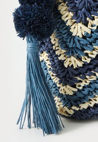 Borsa intrecciata in rattan blu e crema. Presenta pompon morbidi e una frangia testurizzata. Il motivo a zigzag distintivo aggiunge profondità.