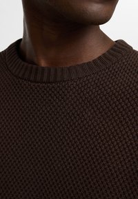 Suéter marrón de punto con un cuello redondo acanalado. La textura presenta un patrón uniforme y ajustado, mostrando un color cálido y rico.