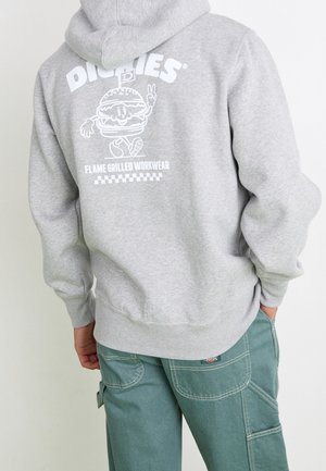 Person trägt einen hellgrauen Hoodie mit einem Cartoon-Burger und dem Text "Dickies Flame Grilled Workwear" auf dem Rücken, kombiniert mit grünen Arbeitshosen.