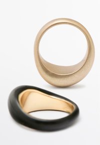 Due anelli esposti; uno è un semplice anello d'oro opaco, l'altro è una fascia nera lucida con un interno dorato, entrambi hanno forme arrotondate.