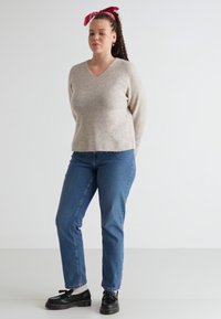 Beige knit sweater with a V-neck, paired with blue straight-leg jeans. Dark lace-up shoes and a red bandana in hair. Smooth texture and casual fit.
