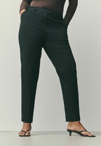 Pantalon noir sur mesure avec une coupe droite, doté de poches latérales et d'une texture lisse, associé à des tongs à talons noires.