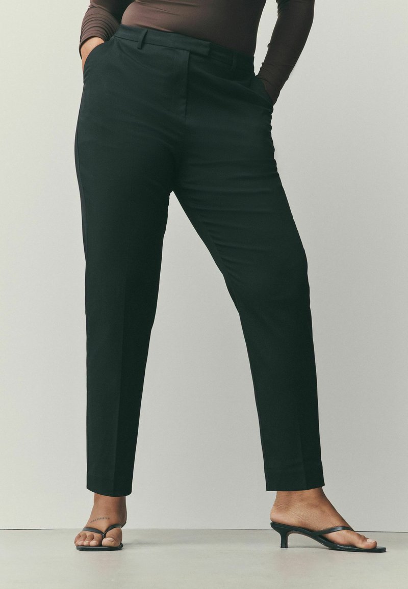 Pantalon noir sur mesure avec une coupe droite, doté de poches latérales et d'une texture lisse, associé à des tongs à talons noires.