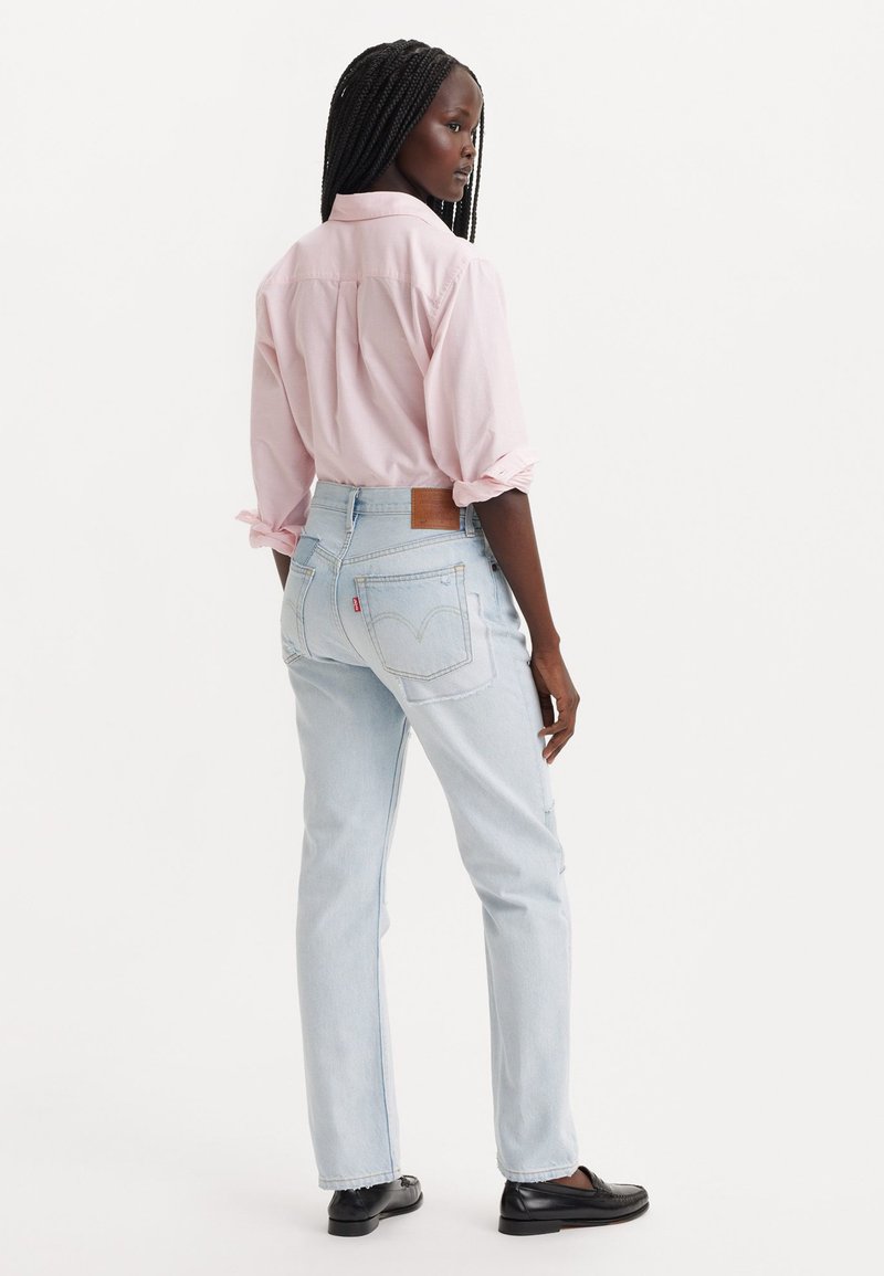 Jeans dritti di colore blu chiaro con una toppa in pelle marrone e un pannello rosso, abbinati a una camicia button-up rosa chiaro, visti da dietro.