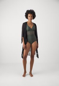 Maillot de bain une pièce vert avec un décolleté en V et une bande texturée ornée d'or. Kimono noir léger avec des franges.