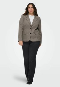 Žena oblečená v šedé kostkované blazer, bílé tričko, černé džíny a černé boty, stojí proti jednobarevnému světlému pozadí.