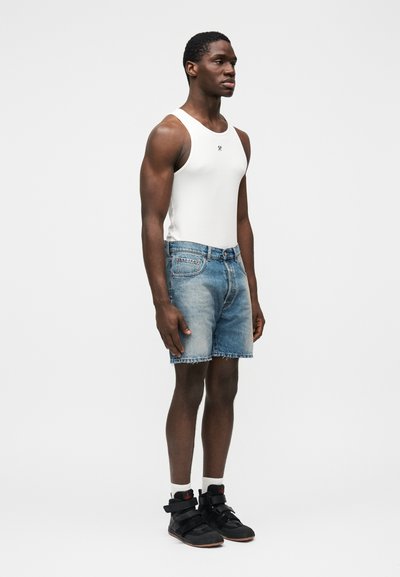 Biele tielko, svetlomodré riflové šortky s roztrhaným lemom. Čierne topánky so zapínaním, biele ponožky. Model stojí tvárou doľava na jednoduchom pozadí.