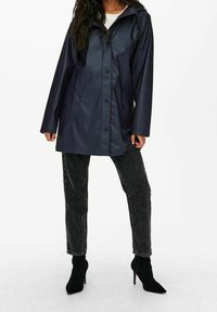 Imperméable marine avec capuche, boutons-pression et deux poches latérales. Porté avec un jean foncé et des bottines noires à talons hauts.