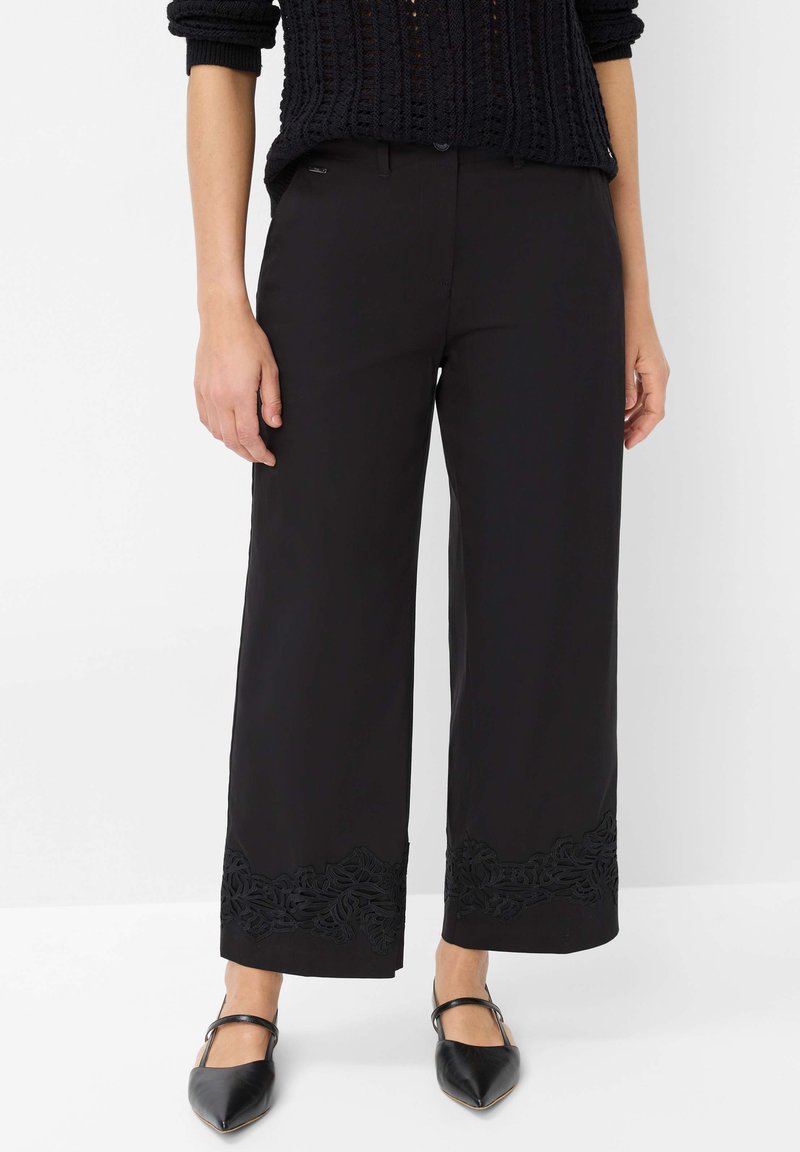 Zwarte broek met wijde pijpen en complexe zwarte bloemenborduursels aan de zoom, gemaakt van zachte stof. Gecombineerd met puntige zwarte schoenen.