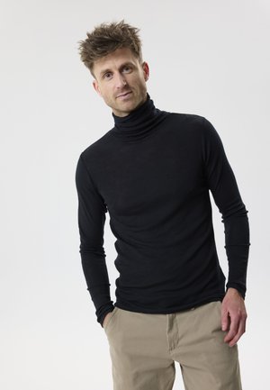 Bărbat cu păr zburlit, purtând un pulover negru cu guler înalt și pantaloni bej, stând cu o mână în buzunar, pe un fundal simplu.