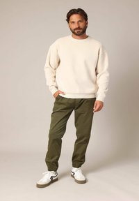 Crème-kleurige gebreide trui met geribde boorden, gecombineerd met olijfgroene tapered broek en witte sneakers met zwarte accenten.