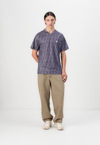 Purple v-neck t-shirt featuring an abstract pattern in darker shades, paired with loose beige pants and light-colored sneakers.