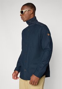 Camicia blu navy con colletto alto e bottoni marroni. Presenta una tasca sul petto e una toppa con logo sulla manica. Tessuto dalla texture liscia.