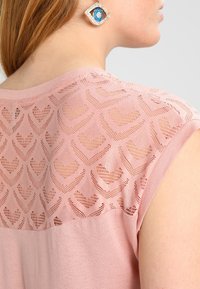 Žena v růžovém bezramenném topu s krajkovým zadním dílem srdíčkového vzoru a visícími náušnicemi ve tvaru oka v modré a stříbrné barvě.