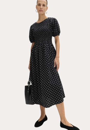 Frau trägt ein schwarzes Midikleid mit weißem Blumenmuster, Puffärmeln, hält eine schwarze Handtasche und trägt schwarze flache Schuhe.