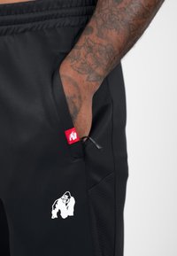 Zwarte sportbroeken met een ritszak, voorzien van een wit gorilla-logo en een rode label met een trekkoord. Gelaagd zijpanelen.