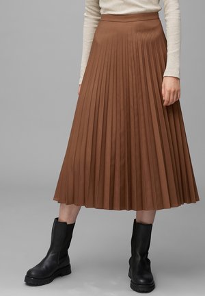 Frau trägt einen braunen plissierten Midirock, beige geripptes Langarmoberteil und schwarze klobige Stiefel, steht vor einem grauen Hintergrund.