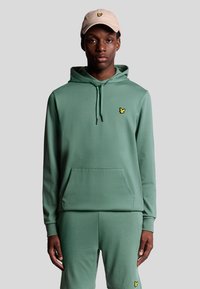 Felpa verde realizzata in un morbido tessuto elastico, con una tasca frontale e un piccolo logo giallo. Abbinata a pantaloni jogger verdi coordinati.