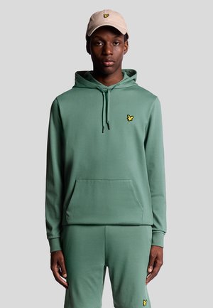 Un sweat à capuche vert en tissu doux et extensible, avec une poche avant et un petit logo jaune. Associé à un pantalon de jogging vert assorti.