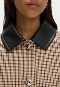 Giacca in tessuto a scacchi marrone e beige con colletto in pelle nera, dotata di un bottone argentato lucido all'apertura del collo.