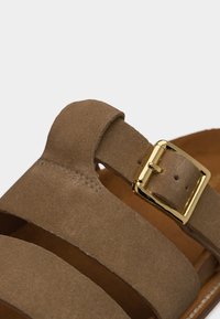 Ρυθμιζόμενο λουράκι σανδαλιού από καφέ σουέντ με αγκράφα από χρυσούς τόνους μετάλλου, που δείχνει λεπτομερή ραφή και οπές για ρύθμιση.