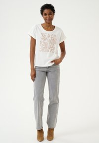 T-shirt blanc à manches courtes avec un motif paisley cuivré sur le devant, associé à un jean en denim gris et des bottines brunes.