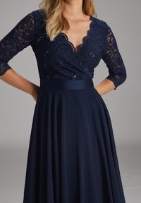 Marineblaues Kleid mit gewelltem Spitzen-Oberteil und dreiviertelärmligen Ärmeln, verziert mit Pailletten. Volles, fließendes Rockteil mit einem Taillengürtel aus Satin.