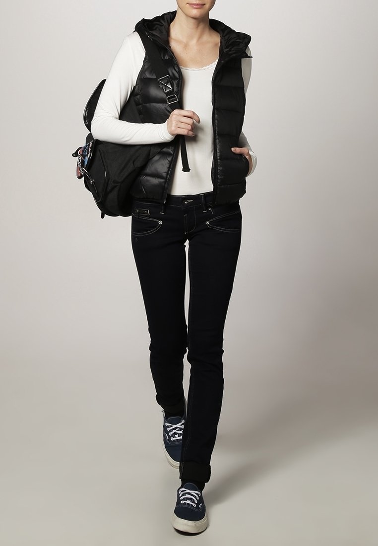Jeune personne portant une doudoune noire sans manches sur une chemise blanche à manches longues, jean noir, baskets bleu marine, portant un sac à dos noir sur une épaule.