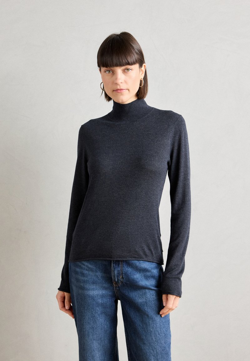 Tettsittende mørkegrå genser med høy hals og lange ermer, laget av mykt materiale, kombinert med blå jeans, som viser et enkelt og rent design.
