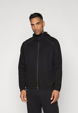 Chaqueta negra con cremallera y tejido texturizado, capucha y bolsillos con cremallera. Presenta un diseño ajustado y detalles contrastantes en los hombros y el collar.
