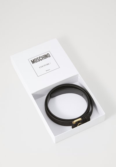 Must mustripaadi valges karbis, millel on kuldse tooniga lukk, sile tekstuur ja minimalistlik disain. Etikett kannab nime "Moschino Couture!"