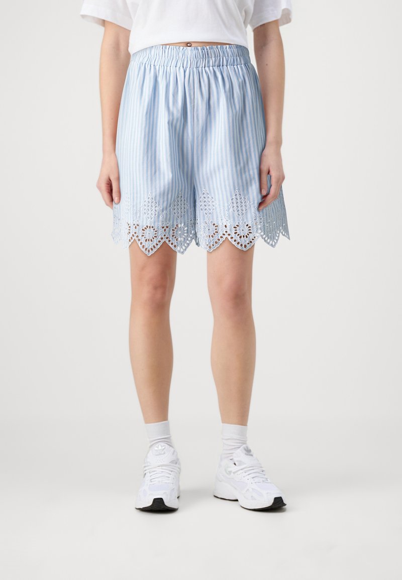 Personne portant un short rayé bleu clair avec une bordure en dentelle blanche, des chaussettes blanches, des baskets blanches et un t-shirt blanc sur un fond uni.