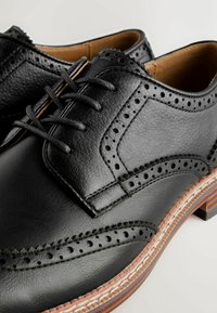 Chaussures habillées brogue en cuir noir avec perforations décoratives, laçage fermé et semelles cousues marron contrastées sur un fond clair.