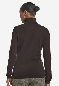 Kobieta z włosami w koksz, nosząca złote kolczyki w kształcie obręczy, ciemnobrązowy sweter z golfem i spodnie w jodełkę, odwrócona plecami.