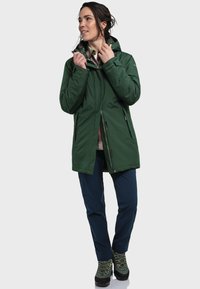 Green insulated jacket with a hood, zippered front, side pockets, and a relaxed fit, paired with navy pants and green hiking boots.