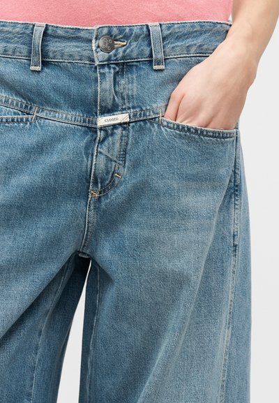 Main dans la poche d'un jean en denim bleu ample avec bouton fermé et étiquette, porté avec un haut rose.