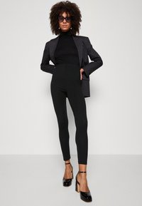 Leggings noires taille haute avec une texture lisse, associées à un col roulé noir ajusté et à un blazer noir à rayures fines. Talons plats noirs.